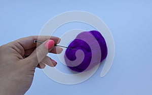 Woman`s hand with pink manicure holding the steel needle above piece of violet wool on white background. Concept of felting creati