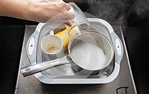 A woman& x27;s hand pours hot water into a basin with dirty dishes and environmentally friendly oxygen bleach. Washing