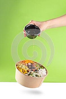 A woman's hand pours soy sauce into a floating bowl of salad poke and tuna, carrot, cucumber, and pineapple with a green