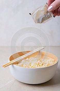 Woman`s hand pours spices from a bag into a bamboo plate bowl with egg noodles on a light background