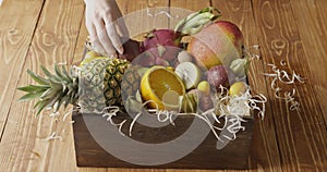 Woman`s hand put wooden box with assortment of exotic fresh organic fruits on a wooden table. Motion, 4K UHD video, 3840