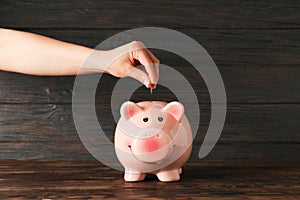 Woman`s hand puts a coin in the happy piggy bank on wood table against wooden background, space for text