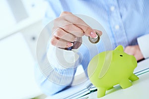 Woman`s hand putting money coin in green piggy bank close-up in office background. Growing business, pension and