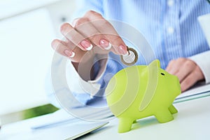 Woman`s hand putting money coin in green piggy bank close-up in office background. Growing business, pension and