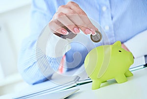 Woman`s hand putting money coin in green piggy bank close-up in office background. Growing business, pension and