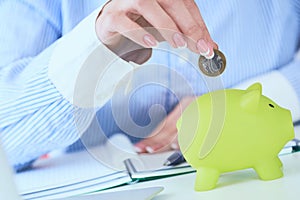 Woman`s hand putting money coin in green piggy bank close-up in office background. Growing business, pension and