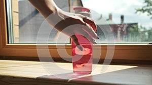 Woman`s hand putting pink plastic bottle of water on windowsill, closeup