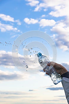 A woman`s hand raises a highly transparent bottle of pure mineral water. Splashes are flying against the background of a blue sky
