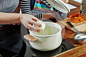 Woman`s hand with raw onion to put it into the boiling water. Opened white saucepan on the black stove. Cooking a soup