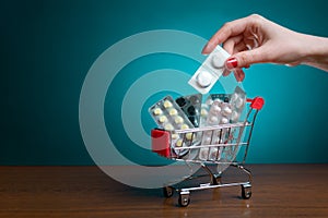 A woman`s hand with a red manicure puts pills in a small cart. Blue background. Copy space. The concept of coronavirus, business