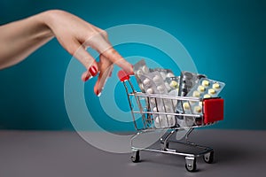 A woman`s hand with a red manicure, rolls with her index finger, a small cart filled with pills and medicines. Blue background.