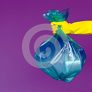 A woman`s hand in a rubber glove holds a transparent green garbage bag with empty plastic bottles.