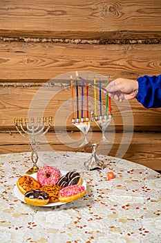 A woman& x27;s hand with a shamash candle lights the Hanukkah candles at the table with donuts