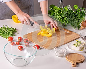 A woman`s hand slices a yellow pepper on a wooden board on a white kitchen table. Preparation of salad