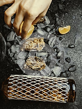 Woman's hand squeezes juice from lemon on oyster shell, dark stone background. Wine bottle and glass. Food aphrodisiac