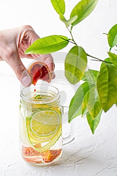 A woman's hand squeezes a slice of red orange into a glass mug with handle with refreshing detox lemonade with sliced