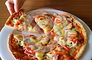 Woman`s hand taking slice of ham and tomato pizza from a white plate
