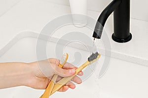 woman's hand washes wooden bamboo toothbrushes from toothpaste after brushing teeth under a stream of water over a sink.