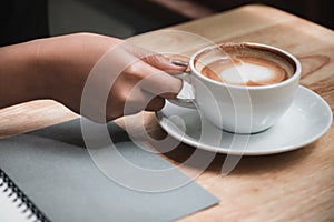 A woman`s hand writing down on a white blank notebook while drinking coffee on wooden table