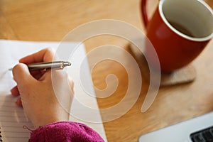 Woman`s hand, writing on a notepad next to a mug of coffe