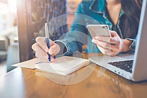 A woman`s hand is writing on a notepad with a pen and a phone wi