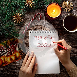 Woman's hand writing Peaceful 2023 on a notepad sheet in winter holiday setting