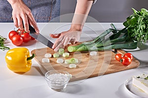 Woman`s hands cut green leek on a wooden board on the kitchen table with various fresh vegetables. Step by Step Salad