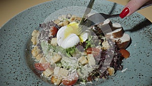 Woman's hands cut a poached egg in Caesar salad with a knife and fork. Close up plate with turkey, tomatoes, lettuce