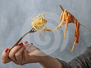 A woman`s hands hold two forks with spaghetti in white on one side and spaghetti with tomato sauce on the other