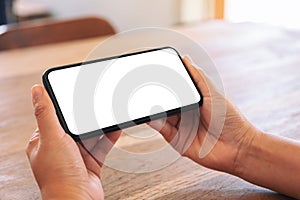 Woman`s hands holding black mobile phone with blank white screen horizontally on wooden table