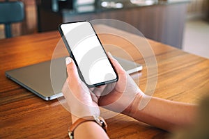 Woman`s hands holding black mobile phone with blank white screen with laptop computer on wooden table