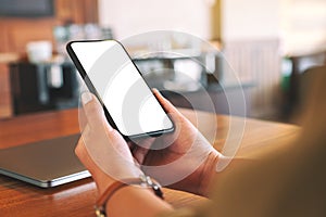 Woman`s hands holding black mobile phone with blank white screen with laptop computer on wooden table