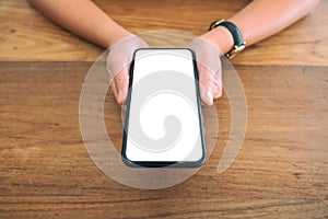 Woman`s hands holding black mobile phone with blank white screen on wooden table
