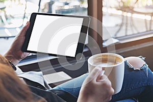 Woman`s hands holding black tablet pc with white blank screen and coffee cup of hot latte sitting in modern cafe