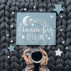 Woman`s hands holding a cup of coffee near a wooden sign with the inscription dream big little one.
