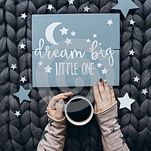 Woman`s hands holding a cup of coffee near a wooden sign with the inscription dream big little one.