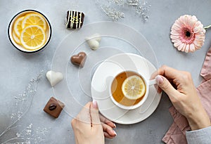 Woman`s hands holding a cup of hot tea with lemon and various sweets on a gray background with flowers.