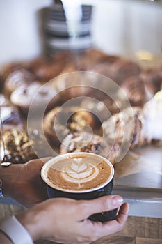 Woman`s hands holding a cup of signature coffee prepared by a barista next to a counter full of croissants. Restaurant. Quiet