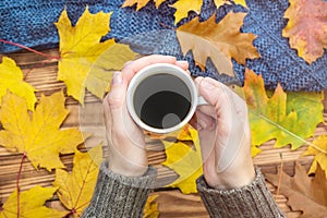 Woman`s hands holding hot cup of coffee over table with autumn leaves and knitted scarf. Top view