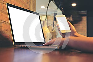 Woman`s hands holding mobile phone while using laptop with blank white screen on vintage wooden table in loft cafe