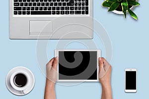 Woman`s hands holding modern tablet over blue background. Office desk with laptop computer, flower, cell phone, tablet and cup of