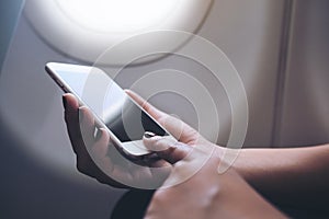 Woman`s hands holding and pointing at a white smart phone with blank black desktop screen next to an airplane window