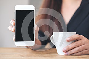 A woman`s hands holding and showing white mobile phone with blank black desktop screen with coffee cups on wooden table