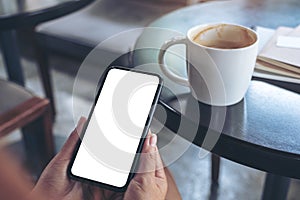 Woman`s hands holding and using a black mobile phone with blank screen with coffee cup on table