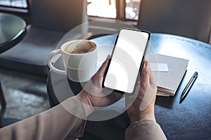 Woman`s hands holding and using a black mobile phone with blank screen for watching with notebook and coffee cup on wooden table