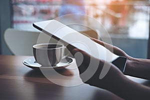 A woman`s hands holding and using tablet pc with coffee cup on wooden table with blur background in cafe