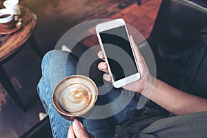 Woman`s hands holding white mobile phone with blank black desktop screen while drinking coffee in cafe