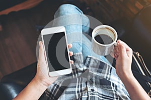 Woman`s hands holding white mobile phone with blank black desktop screen while drinking coffee in cafe