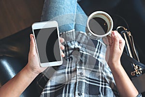 Woman`s hands holding white mobile phone with blank black desktop screen while drinking coffee in cafe