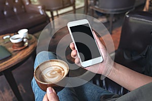 Woman`s hands holding white mobile phone with blank black desktop screen while drinking coffee in cafe